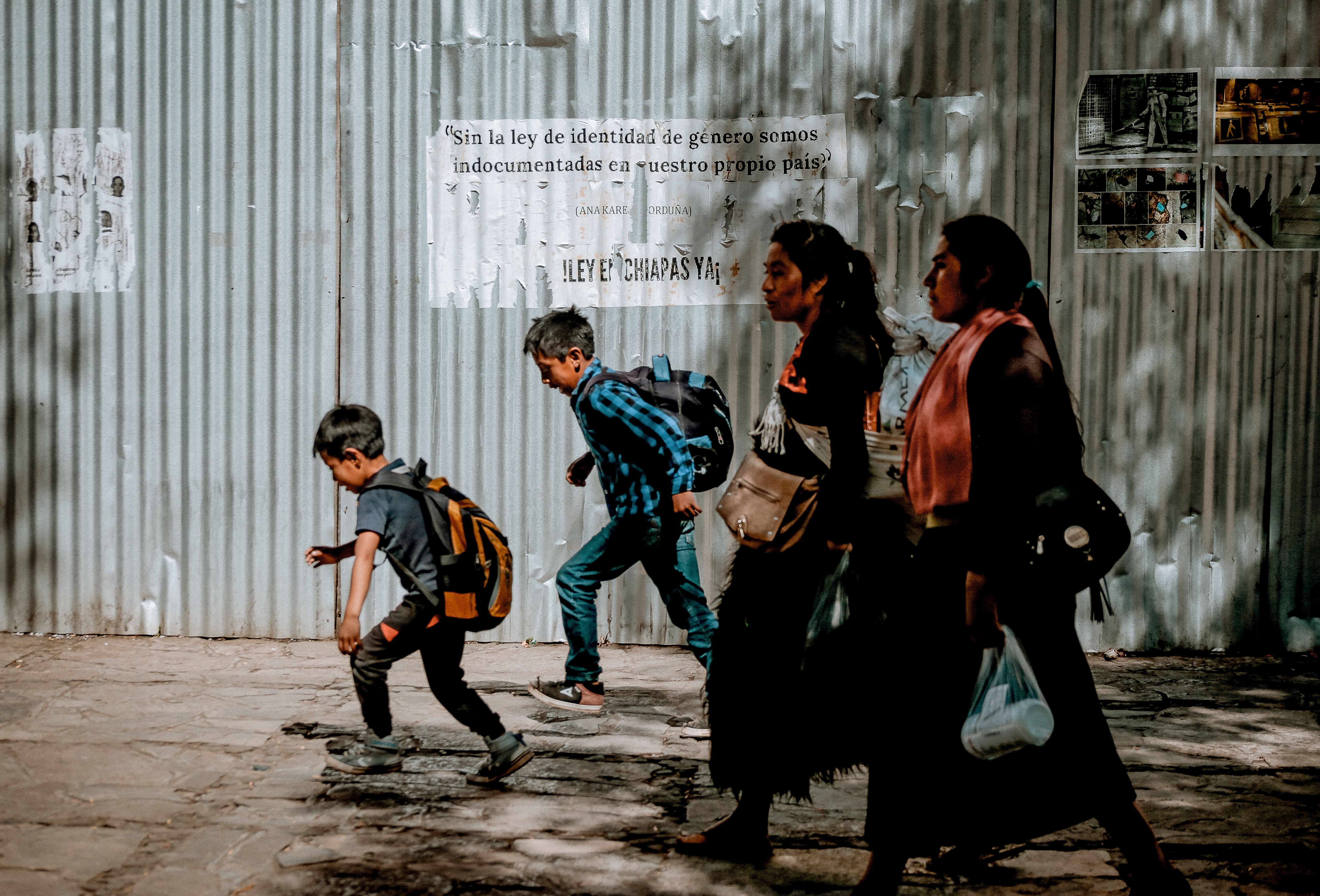 Dos mujeres y dos niños caminan por una banqueta frente a una pared de lámina con carteles informativos.